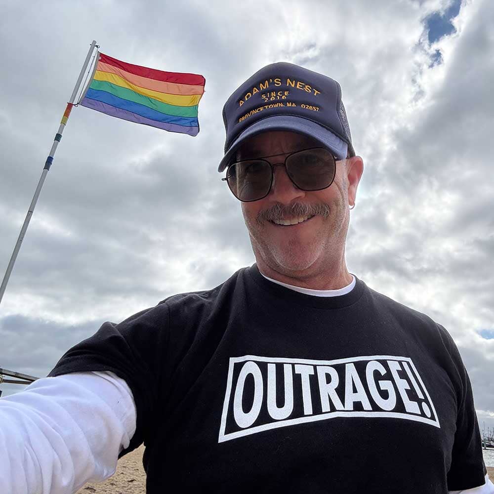 ADAM SINGER wearing a black 'OUTRAGE!' shirt with a rainbow flag in the background