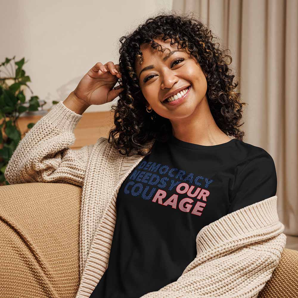 Woman wearing a black t-shirt with "democracy needs your courage" sitting on a couch.