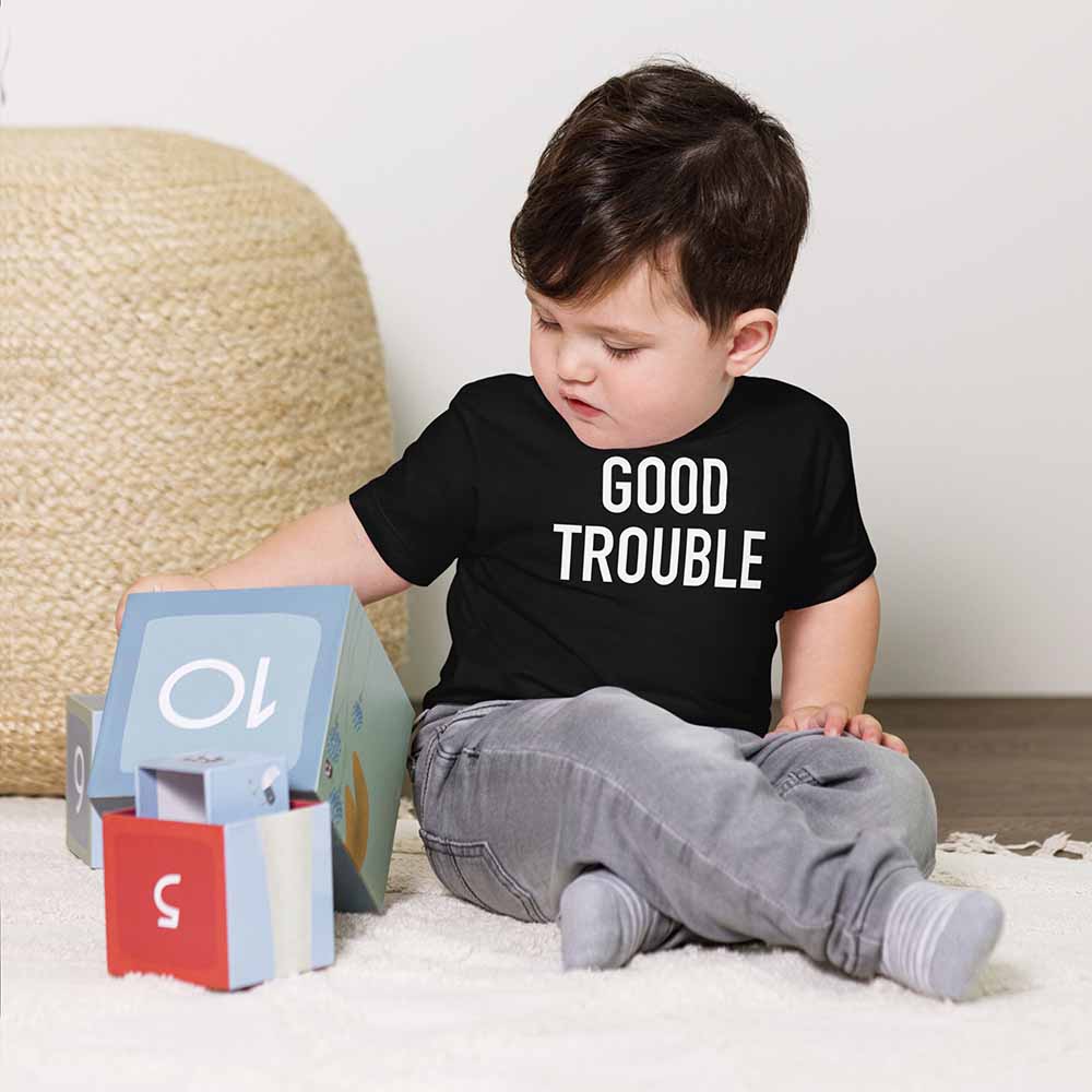 Child wearing a black t-shirt with 'GOOD TROUBLE' text, sitting on a carpeted floor.
