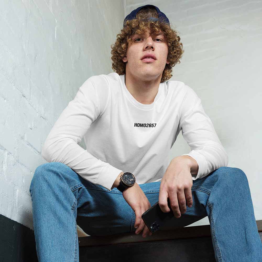 Person wearing a white long-sleeve shirt with HOMO2657 text, blue jeans, and a cap, sitting against a white brick wall.