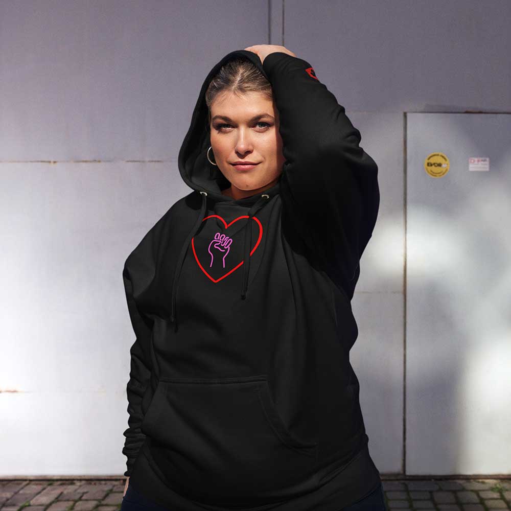 Woman wearing a black hoodie with a heart and solidarity fist design, standing against a plain wall.