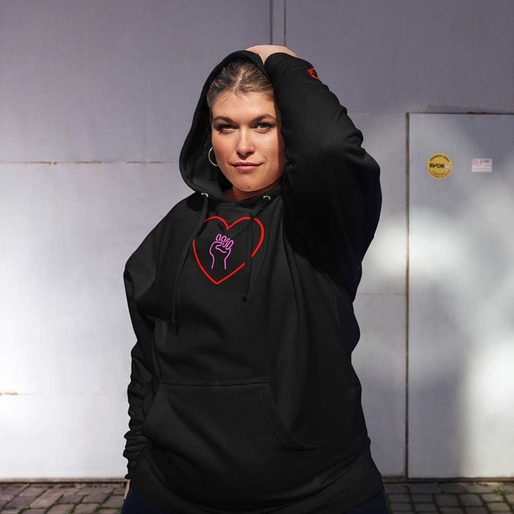 Woman wearing a black hoodie with a heart and solidarity fist design, standing against a plain wall.