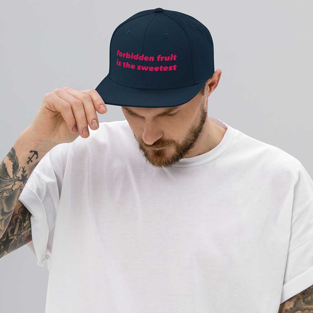 Man wearing a navy flatbill cap with pink text reading forbidden fruit is the sweetestt on a plain background