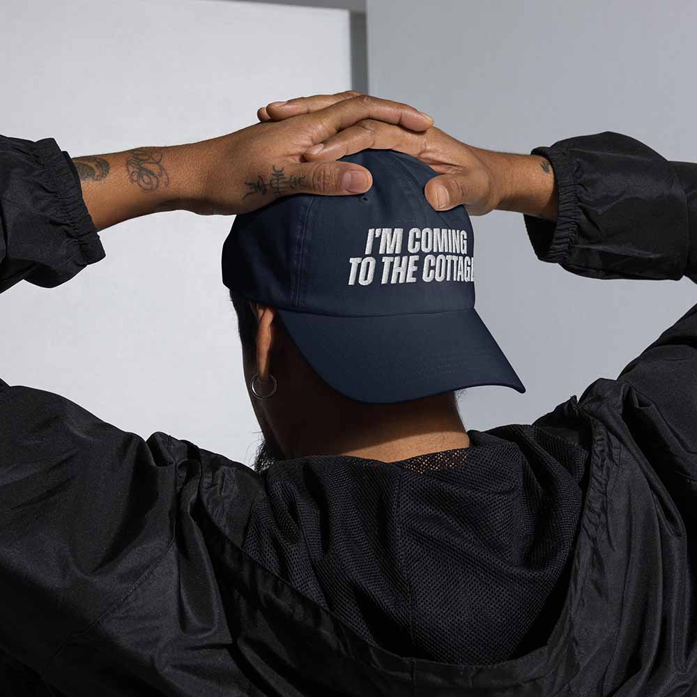 Person wearing a navy blue cap with 'I'm coming to the cottage' text, hands clasped together.