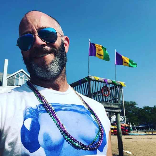 Man with sunglasses and beads standing in front of a deck with flags on a beach.