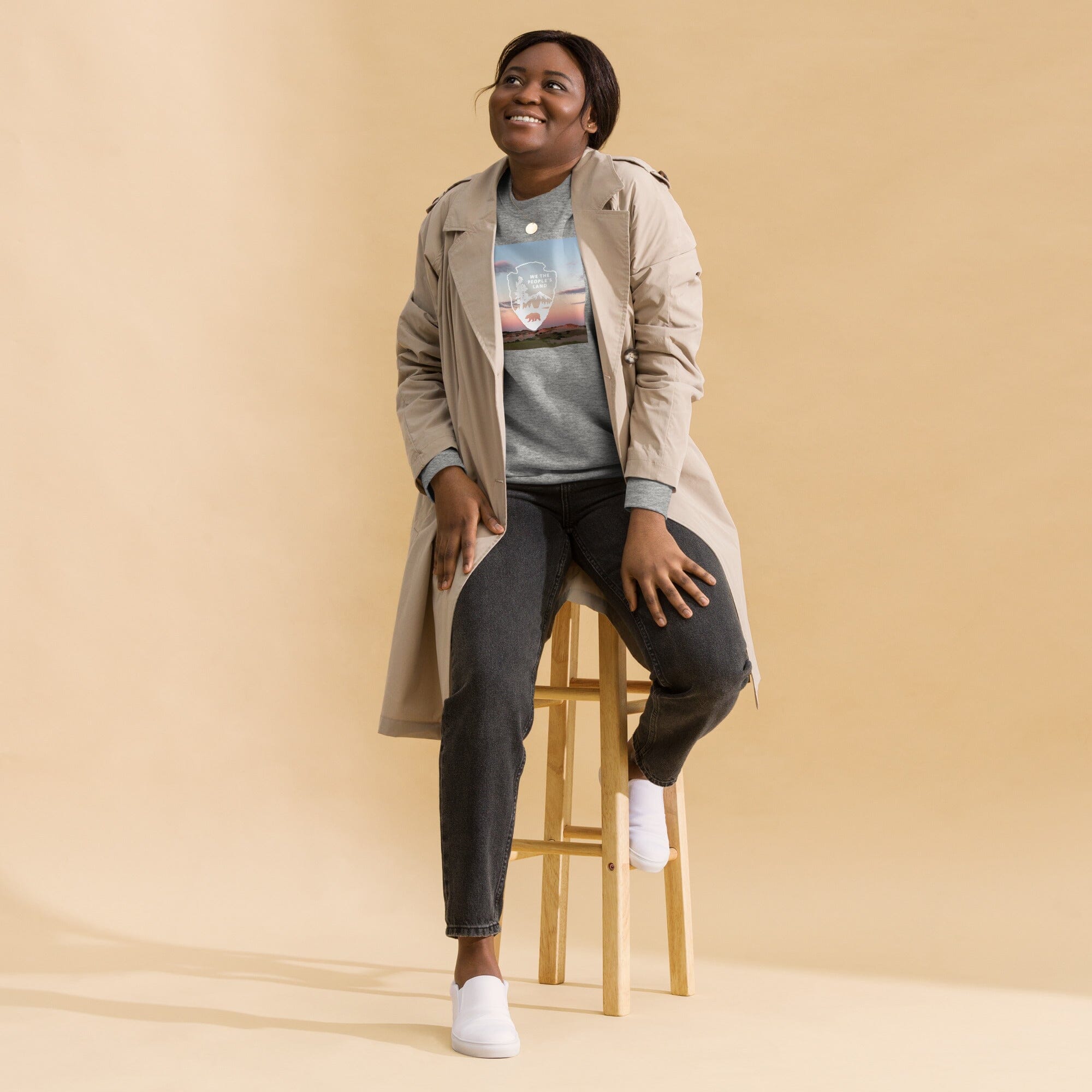 Woman sitting on a stool wearing a beige trench coat, gray shirt, and dark pants against a beige background